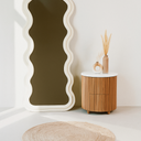Decorative mirror with wavy frame next to a wooden side table with a vase on a white floor.