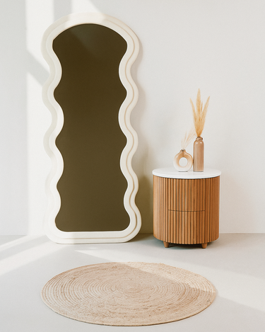 Decorative mirror with wavy frame next to a wooden side table with a vase on a white floor.